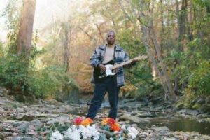 A black man poses with a guitar outside by a creek