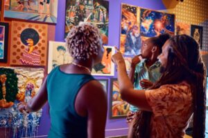 A Black man with dreads holds a little girl up to look at colorful paintings. A black woman with cropped blonde hair stands to his left.