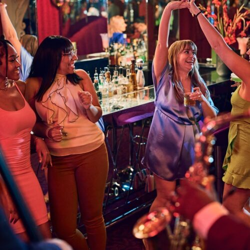 Women dressed in cocktail dresses sip drinks and dance to jazz musicians