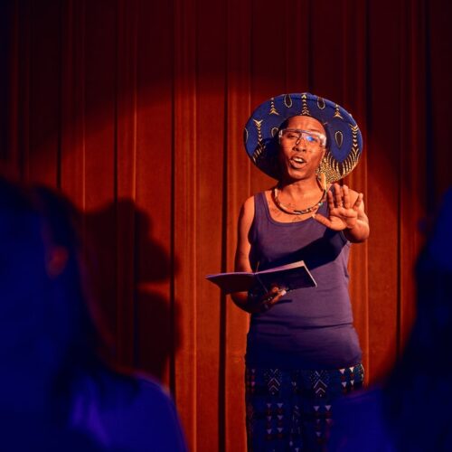 A Black woman with a wide hat performs a spoken word on a stage illuminated in red light
