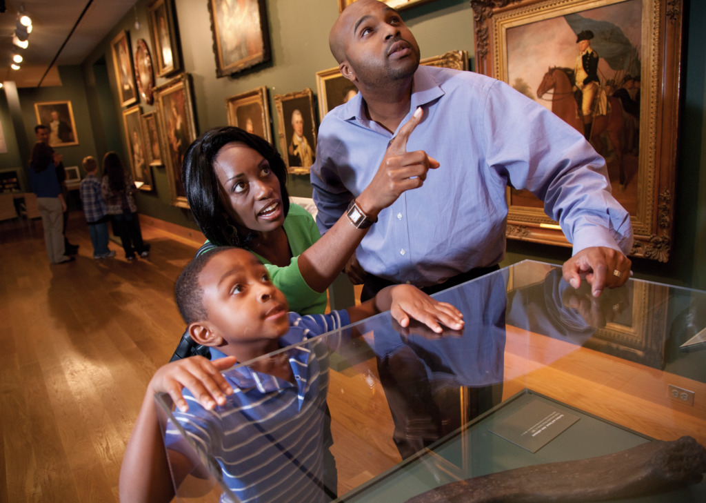 A man, woman and child look at a piece of art in a museum, not visible to the viewer of the photo. The woman points up with a curious expression.