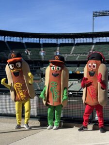 Hot dog mascots representing mustard, relish and ketchup (from left to right).