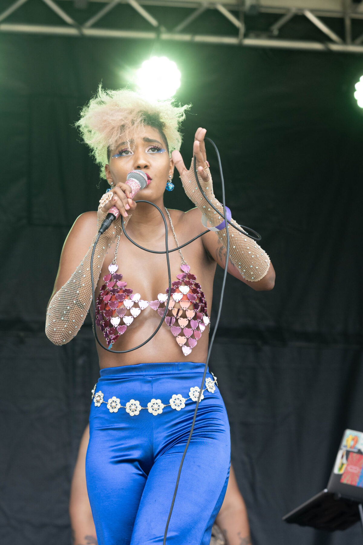 A black female performer with short blonde hair sings into a microphone. She is wearing blue flare pants